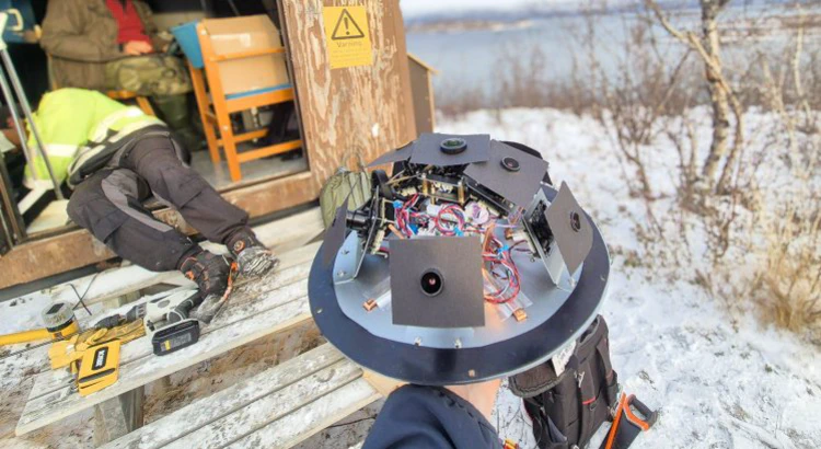 Meteorkamera utan sitt skyddande hölje. I bakgrunden mätstation i vintrigt Abisko med öppen dörr, mängder av verktyg och två av IRFs medarbetare i arbete.